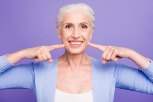 Concept of having strong healthy white perfect teeth at old age. Portrait of old lady with beaming smile pointing on her teeth, isolated over violet background